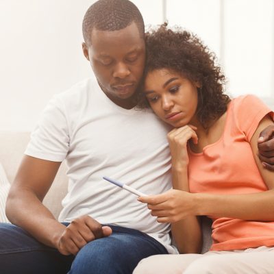 Sad african-american couple after negative pregnancy test result, sitting on couch at home, copy space